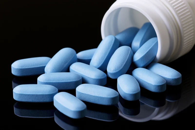 Blue oval pills spilling from a white plastic bottle onto a reflective surface