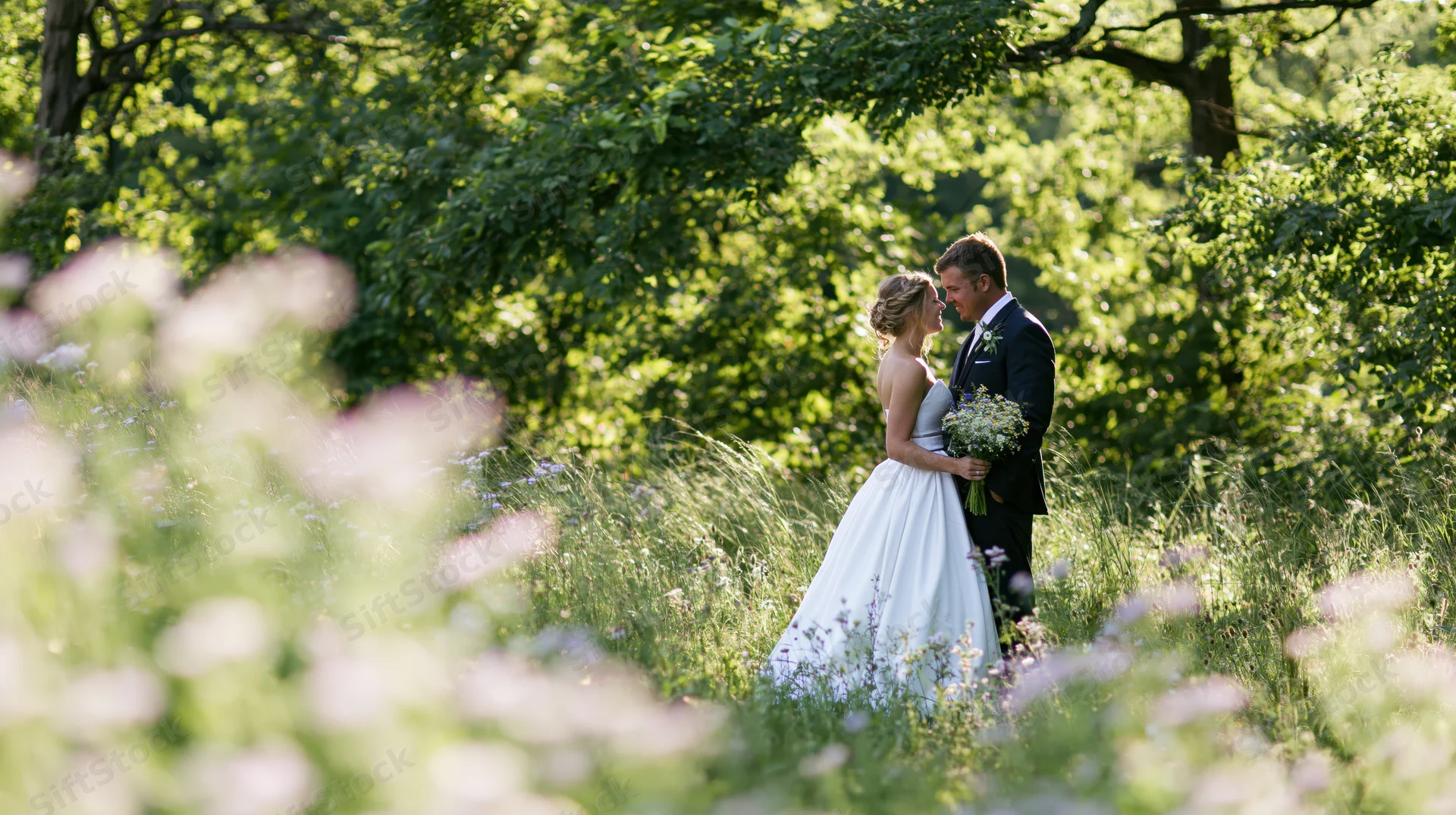 Bride and groom standing close together in a sunlit garden surrounded by greenery and wildflowers