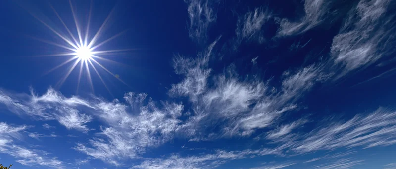 Bright midday sun with starburst lens flare and wispy white clouds in a deep blue sky
