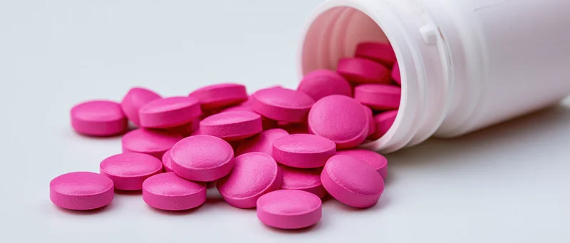 Bright pink round tablets spilling from an overturned white bottle on a plain surface