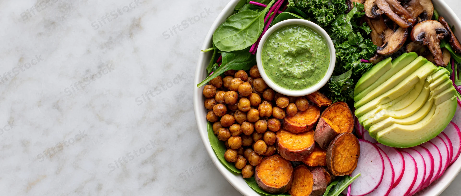 Buddha bowl with roasted chickpeas, sliced avocado, sweet potato, greens, radish, mushrooms, and green dressing on white marble surface