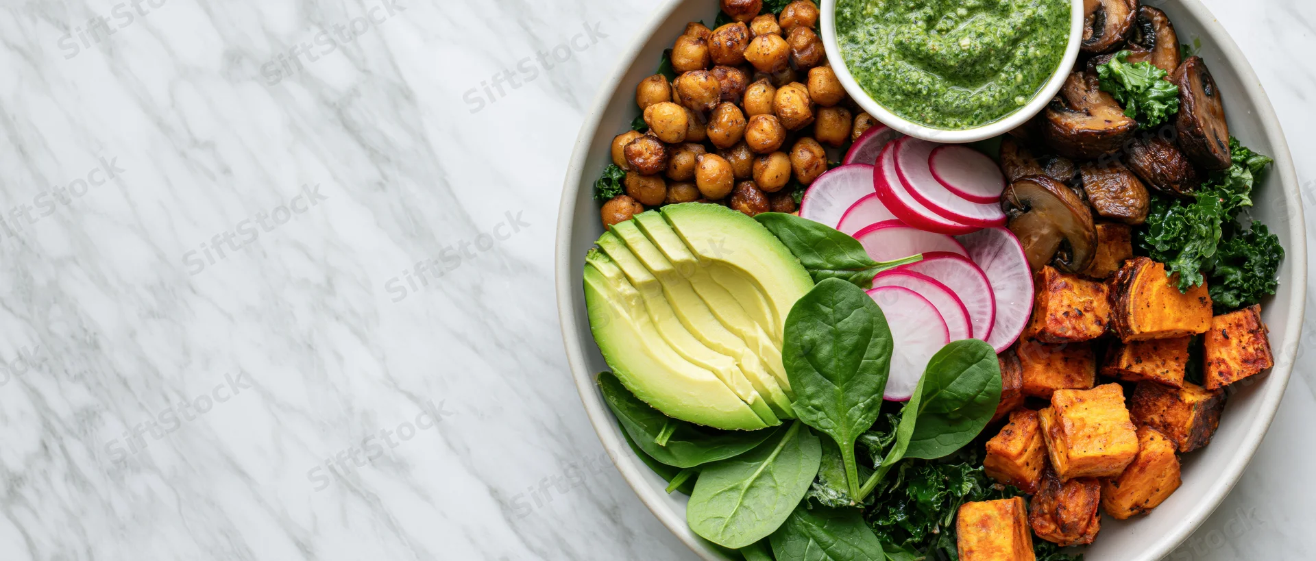 Buddha bowl with roasted chickpeas, sliced avocado, sweet potato, radish, kale, spinach, mushrooms, and pesto on marble countertop