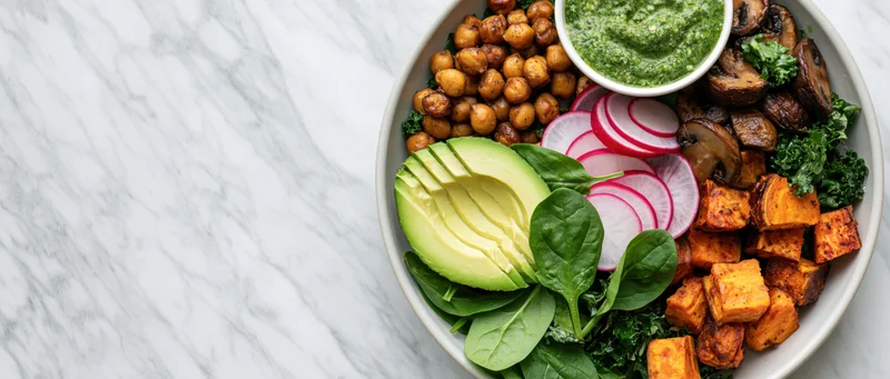 Buddha bowl with roasted chickpeas, sliced avocado, sweet potato, radish, kale, spinach, mushrooms, and pesto on marble countertop