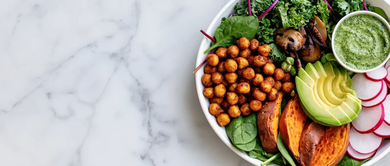 Buddha bowl with roasted chickpeas, sliced avocado, sweet potato, kale, and green dressing on a marble surface