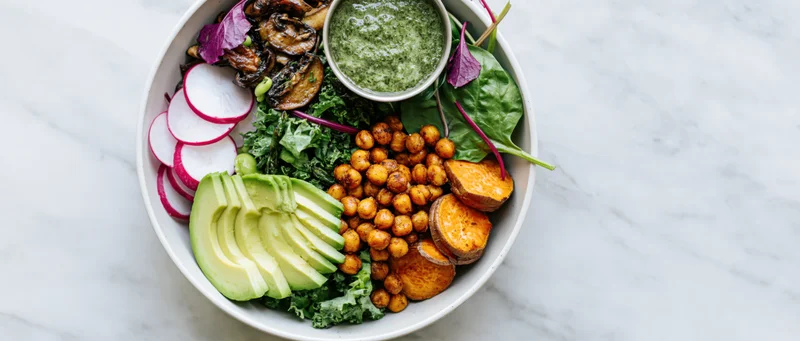 Buddha bowl with roasted chickpeas sliced avocado sweet potato kale and pesto sauce on marble surface