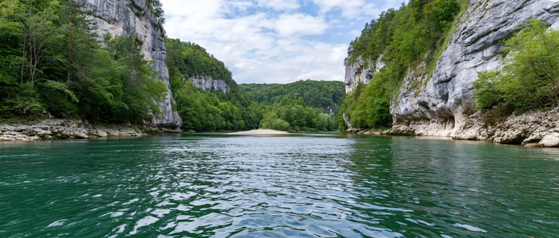 Calm river flowing through a narrow canyon flanked by steep limestone cliffs and dense green forest under a partly cloudy sky
