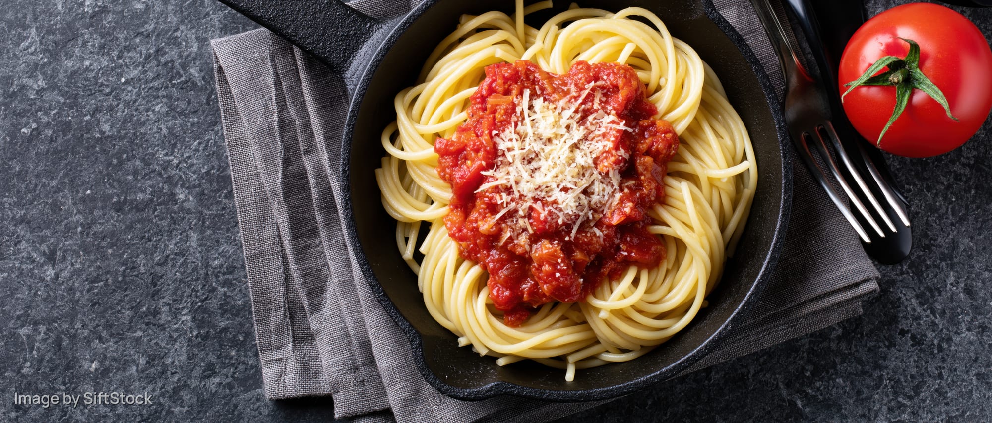 Spaghetti with tomato sauce and cheese in a cast iron skillet on a dark stone surface with a fork and napkin
