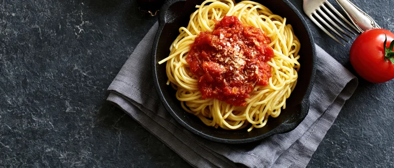 Spaghetti with tomato sauce in a cast iron skillet on a dark surface with a fork and tomato beside it