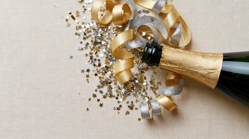 Champagne bottle lying on its side with gold and silver ribbons and star-shaped confetti on a beige linen surface