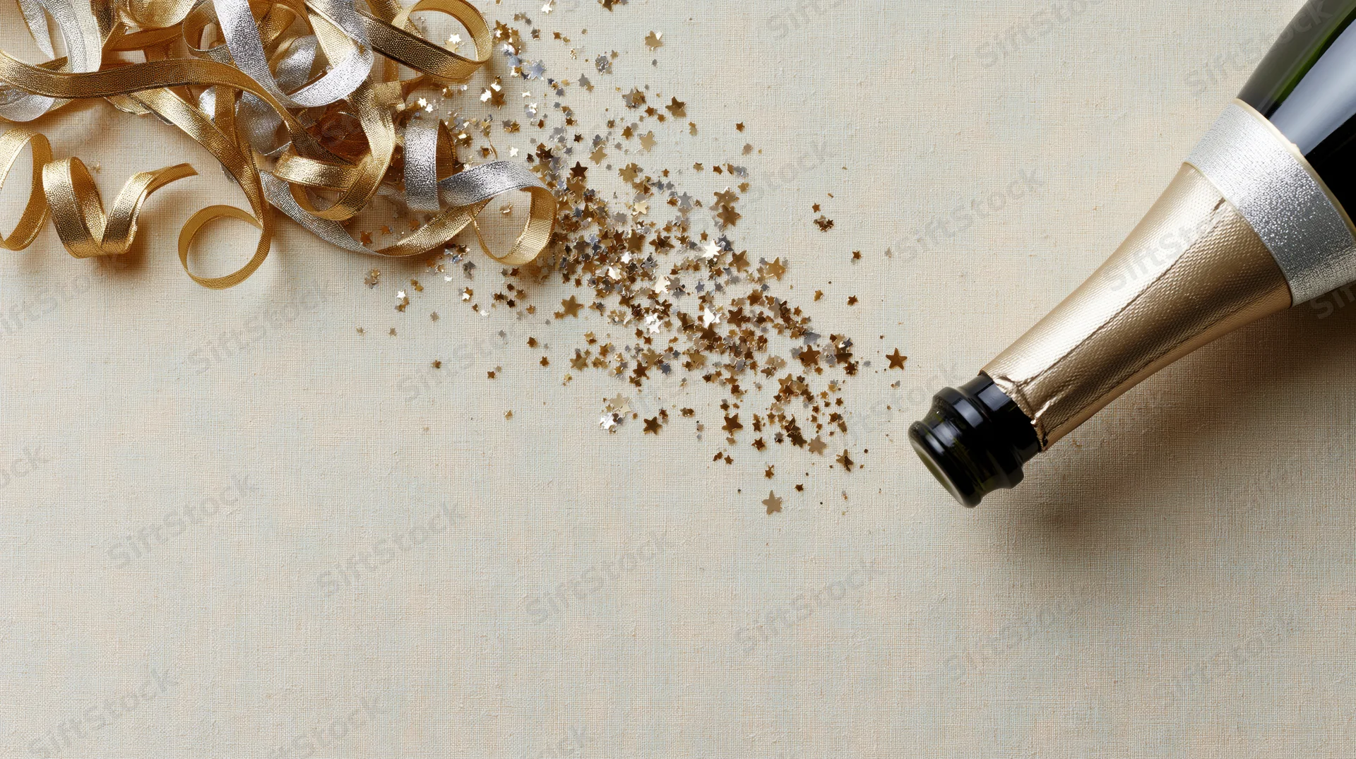Champagne bottle lying on its side with gold and silver ribbons and star-shaped glitter scattered nearby on a light fabric surface