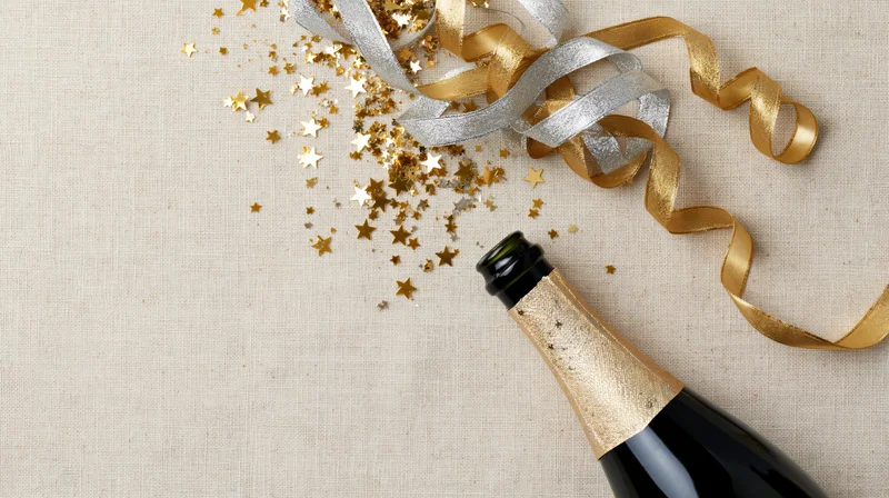 Champagne bottle lying on its side with gold and silver ribbons and star-shaped confetti on a beige linen surface