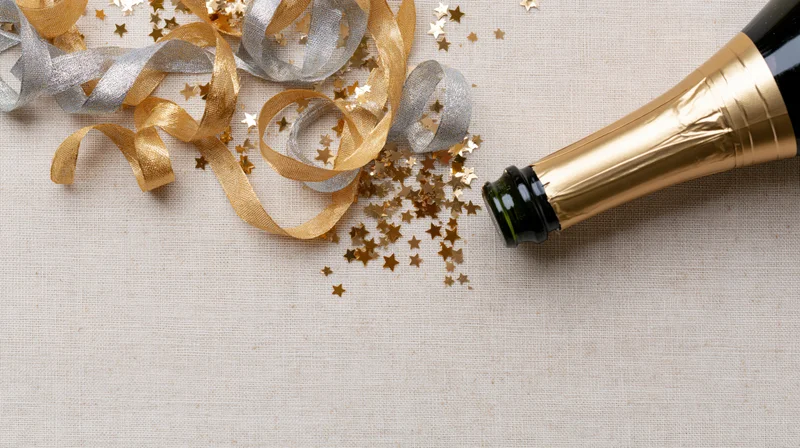 Champagne bottle lying on its side with gold and silver ribbons and star confetti on a linen surface