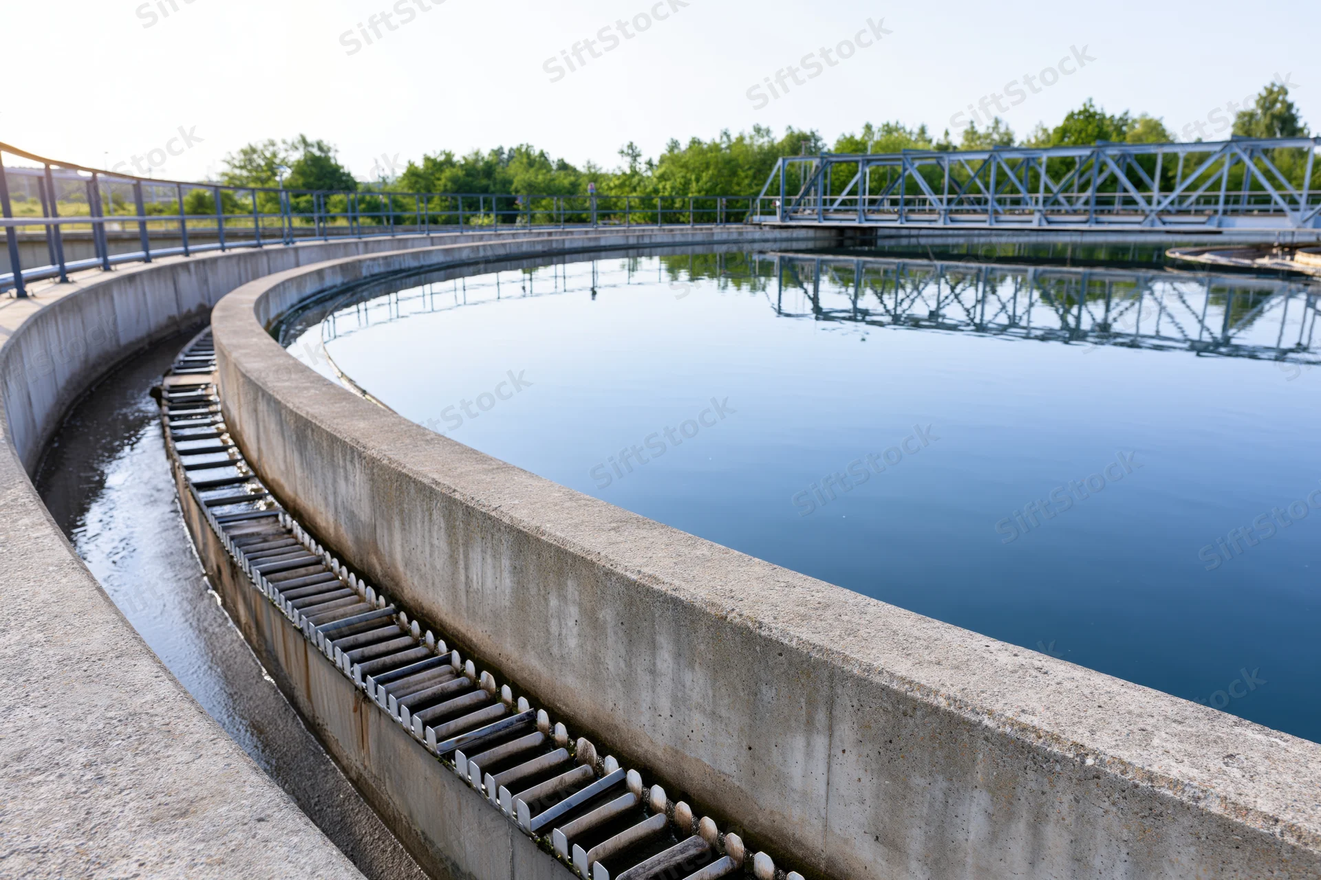 Circular clarifier tank in a municipal wastewater treatment facility with metal bridge and surrounding greenery
