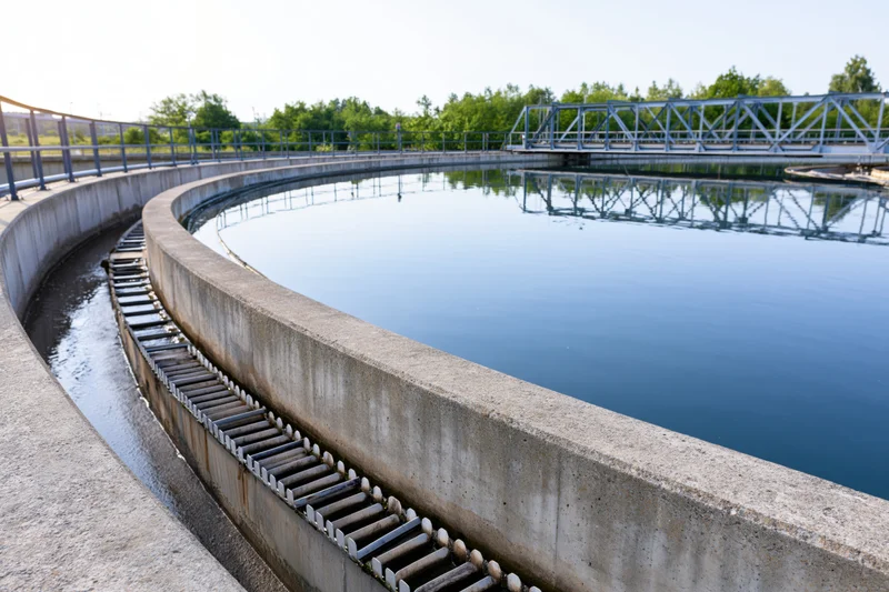 Circular clarifier tank in a municipal wastewater treatment facility with metal bridge and surrounding greenery