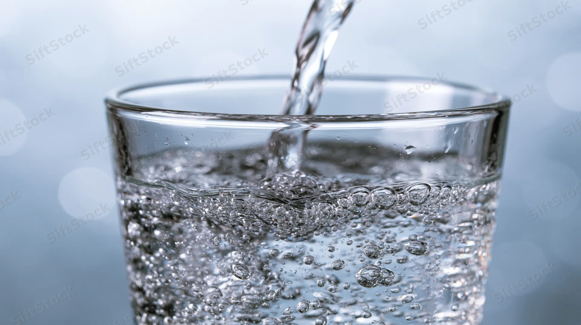 Water being poured into a clear glass, creating bubbles and ripples, on a light blue background