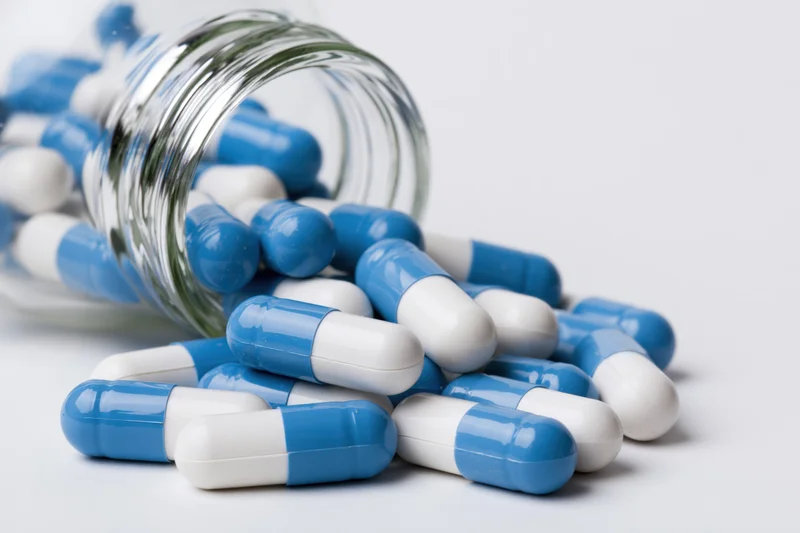 Blue and white capsules spilled from a clear glass bottle on white surface