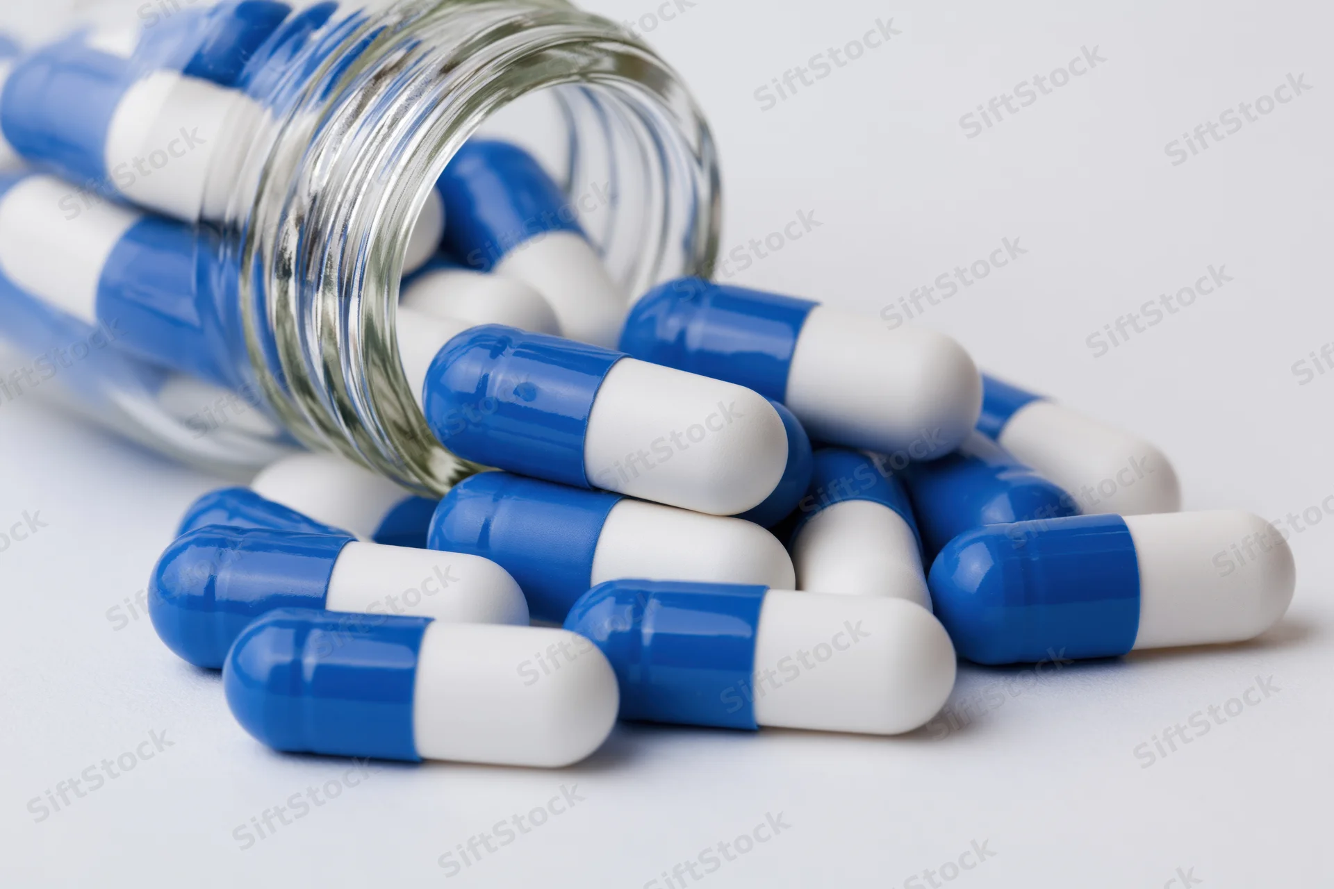 Blue and white capsules spilling from a clear glass bottle on a white surface