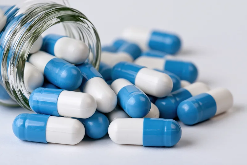 Blue and white capsules spilling from a clear glass bottle onto a white surface