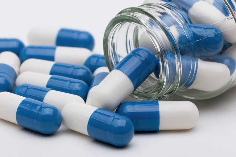 Blue and white capsules spilling from a clear glass bottle on a white surface
