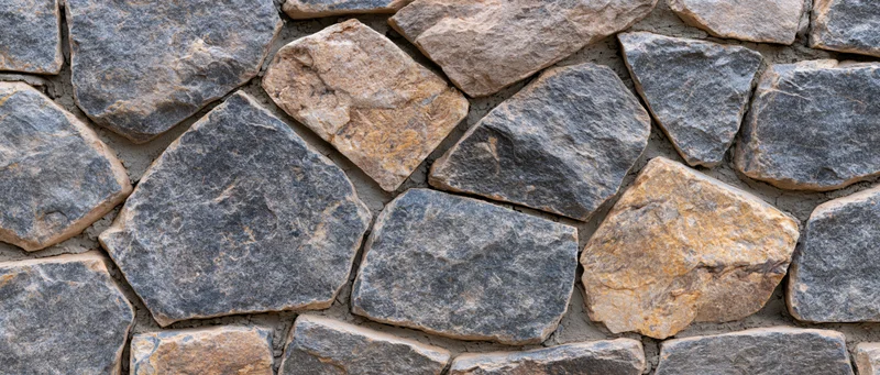 Close-up of a dry-stacked rubble stone wall with irregular gray and beige stones forming a textured exterior surface
