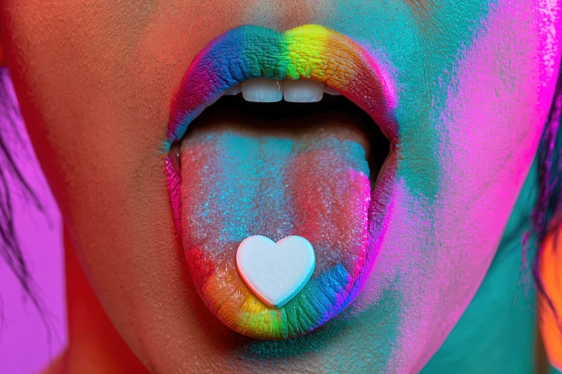 Woman with open mouth and extended tongue holding a heart-shaped candy, face painted in rainbow colors