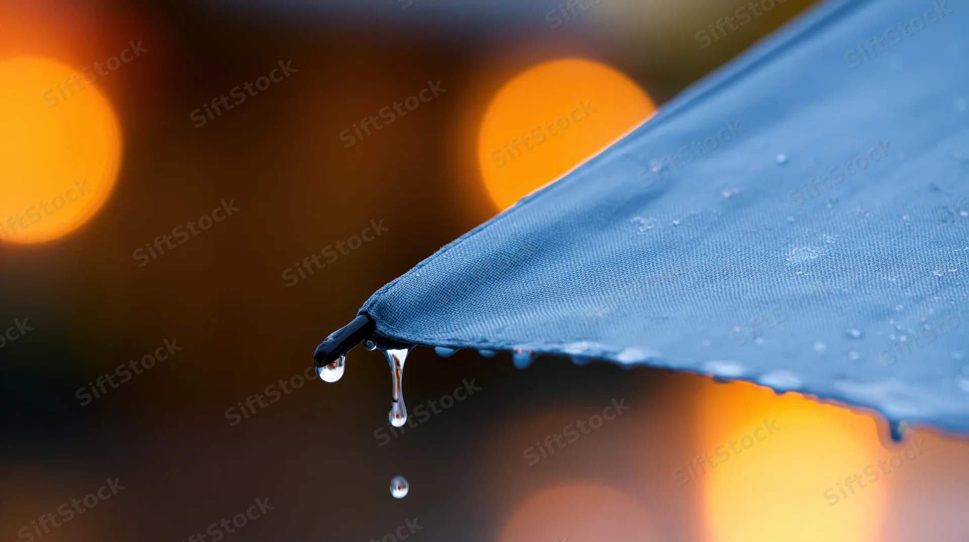 Close-up of rain droplets falling from the edge of a blue umbrella at night with blurred city lights in the background