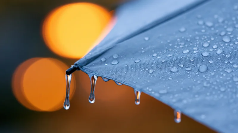 Close-up of rain droplets dripping from the edge of a blue umbrella at night with blurred city lights in the background