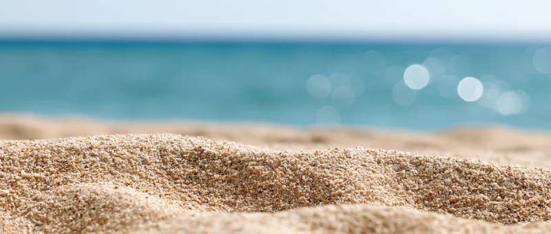 Close-up of sun-warmed beach sand in foreground with blurred ocean and sky in background