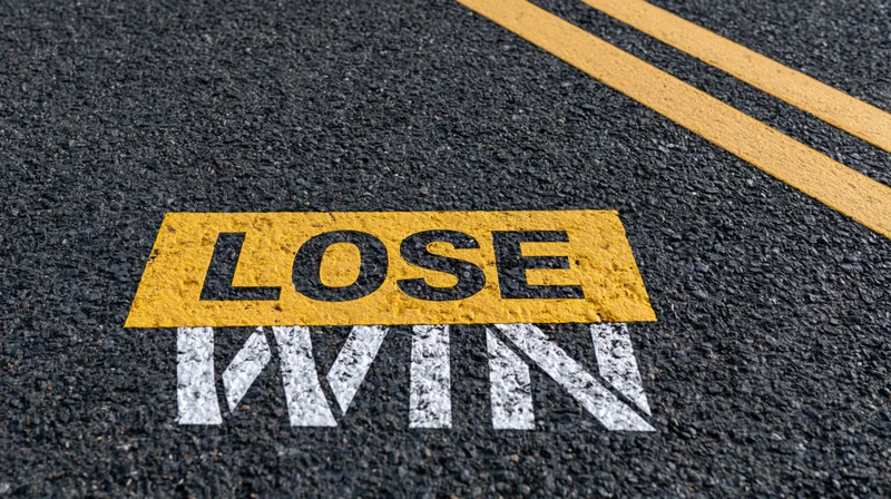 Close-up overhead view of asphalt road with yellow painted stripe covering the word "win" and revealing "lose" beneath