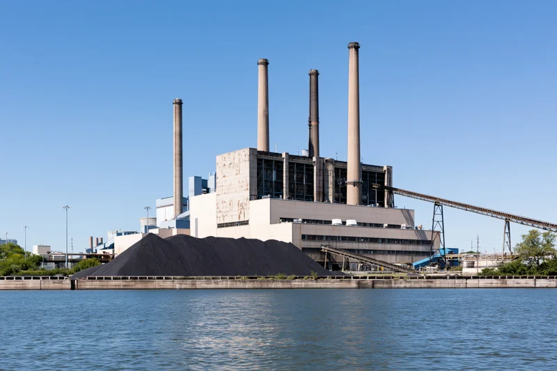 Coal-fired power plant with large coal stockpile beside a river under clear blue sky