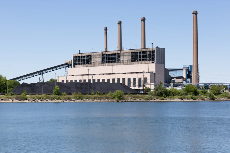 Coal-fired power plant with large coal stockpile beside a river under clear blue sky