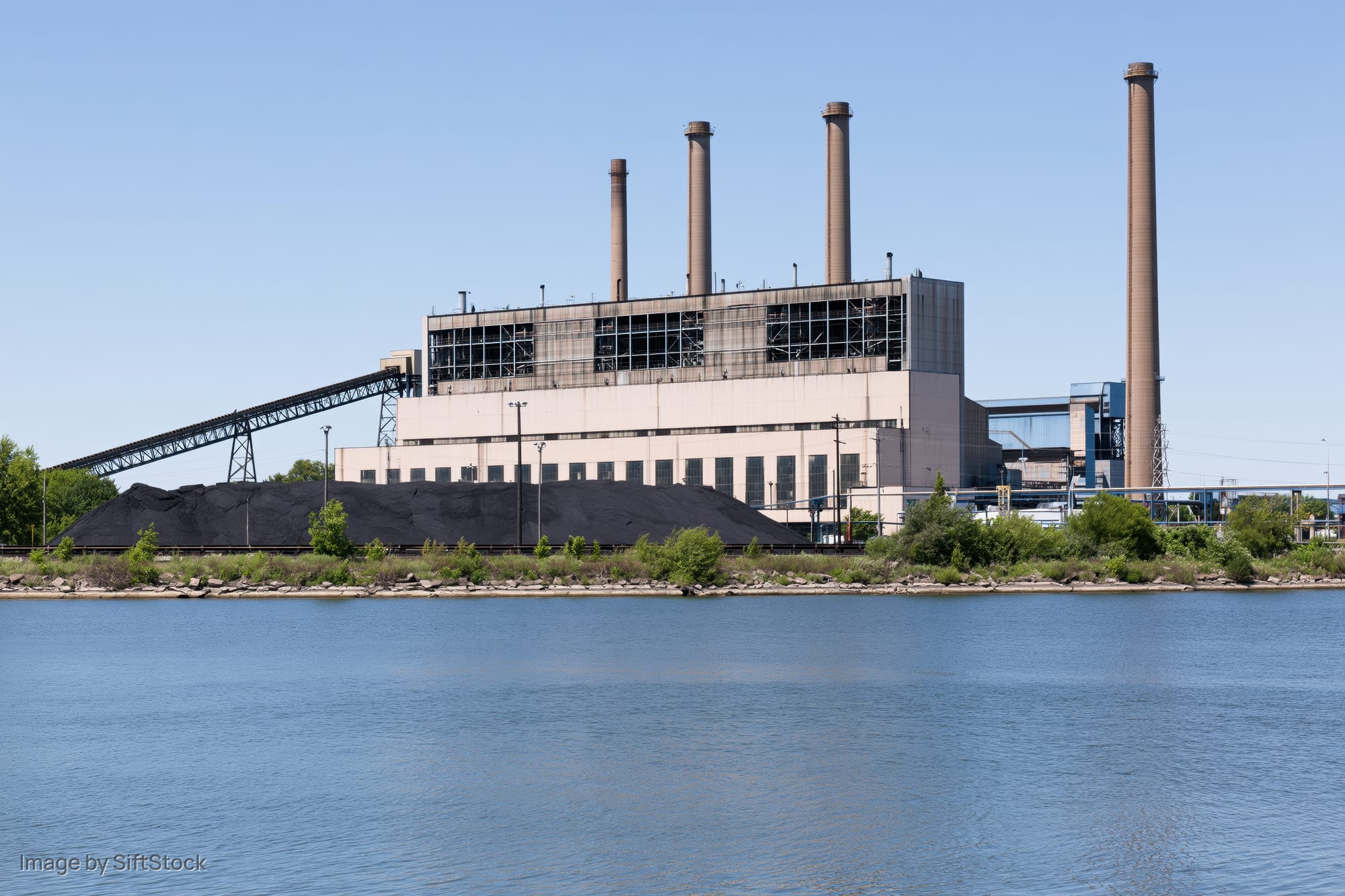 Coal-fired power plant with large coal stockpile beside a river under clear blue sky