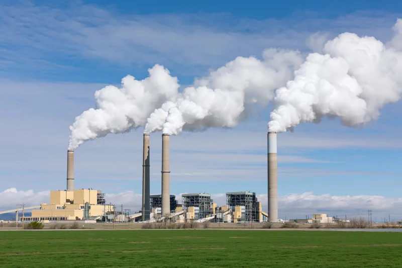 Coal-fired power plant with three tall smokestacks emitting white smoke against a blue sky over a green field