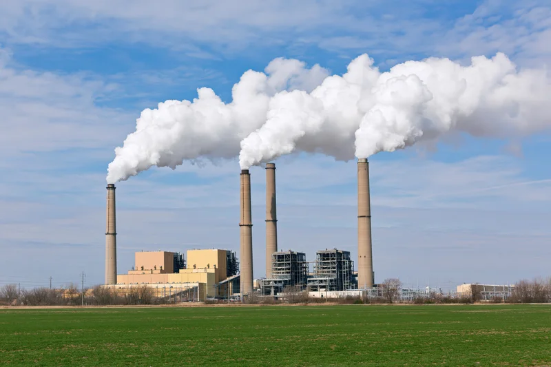 Coal-fired power plant with three tall smokestacks emitting white steam against a blue sky, viewed from a green field