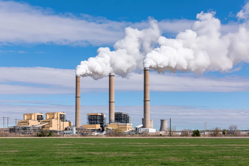 Coal-fired power plant with three tall smokestacks emitting white smoke against a blue sky over a green field