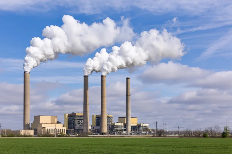Coal-fired power plant with four smokestacks emitting white smoke against a blue sky over a green field