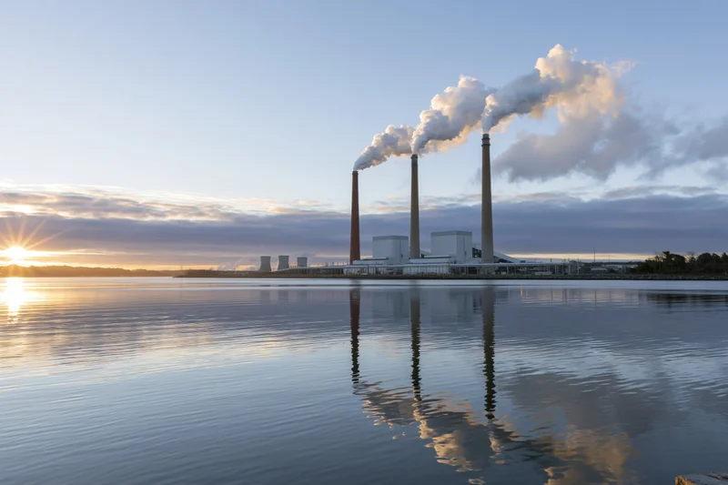 Coal-fired power plant with three smokestacks emitting steam over a calm river at sunset
