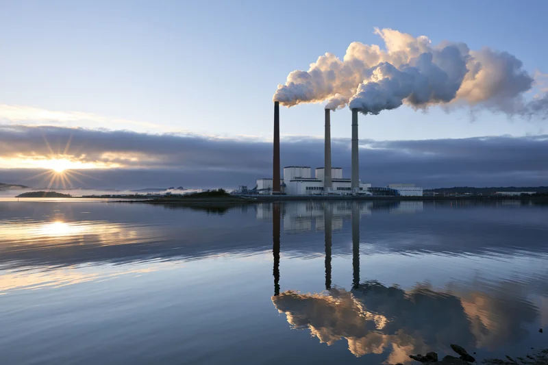 Coal-fired power plant with three smokestacks emitting steam at sunset over calm water