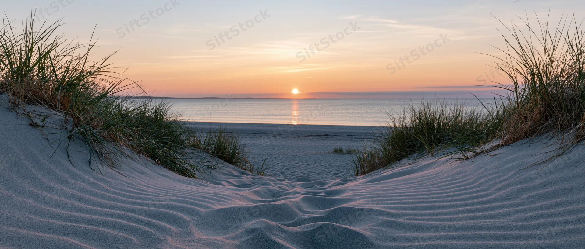 Sand dunes with marram grass framing a view of the ocean at sunset
