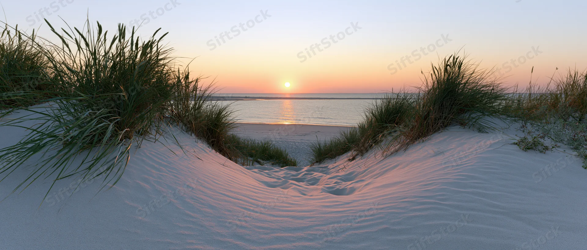 Sand dunes with marram grass framing a view of the ocean at sunset