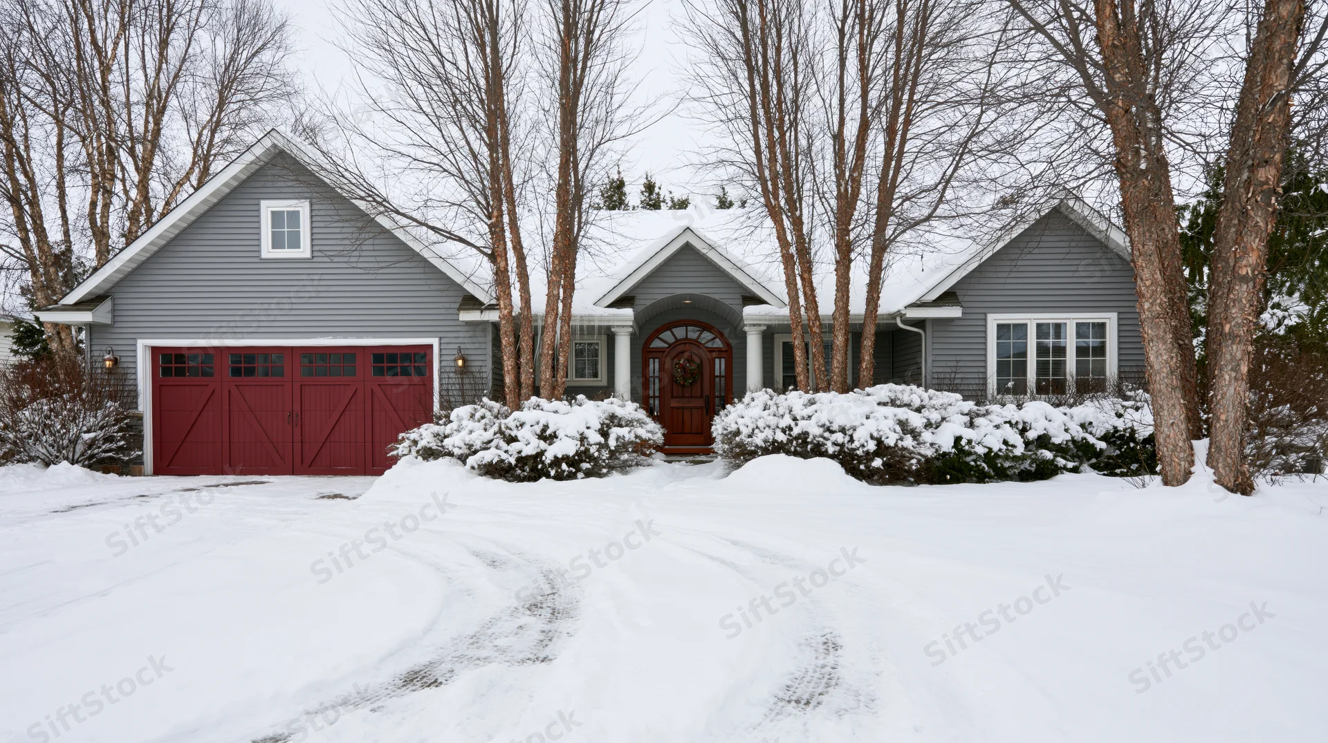 Craftsman-style suburban house with grey vinyl siding and red garage doors in snow-covered yard