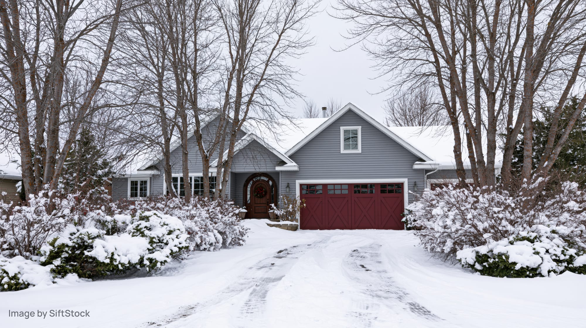 Craftsman-style suburban house with grey vinyl siding and red garage door in snow-covered yard