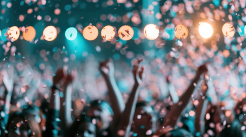 Crowd celebrating with arms raised at indoor event under stage lights and falling confetti