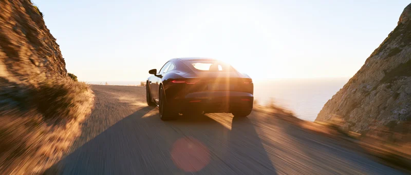 Black sports coupe driving away on narrow coastal road at sunset