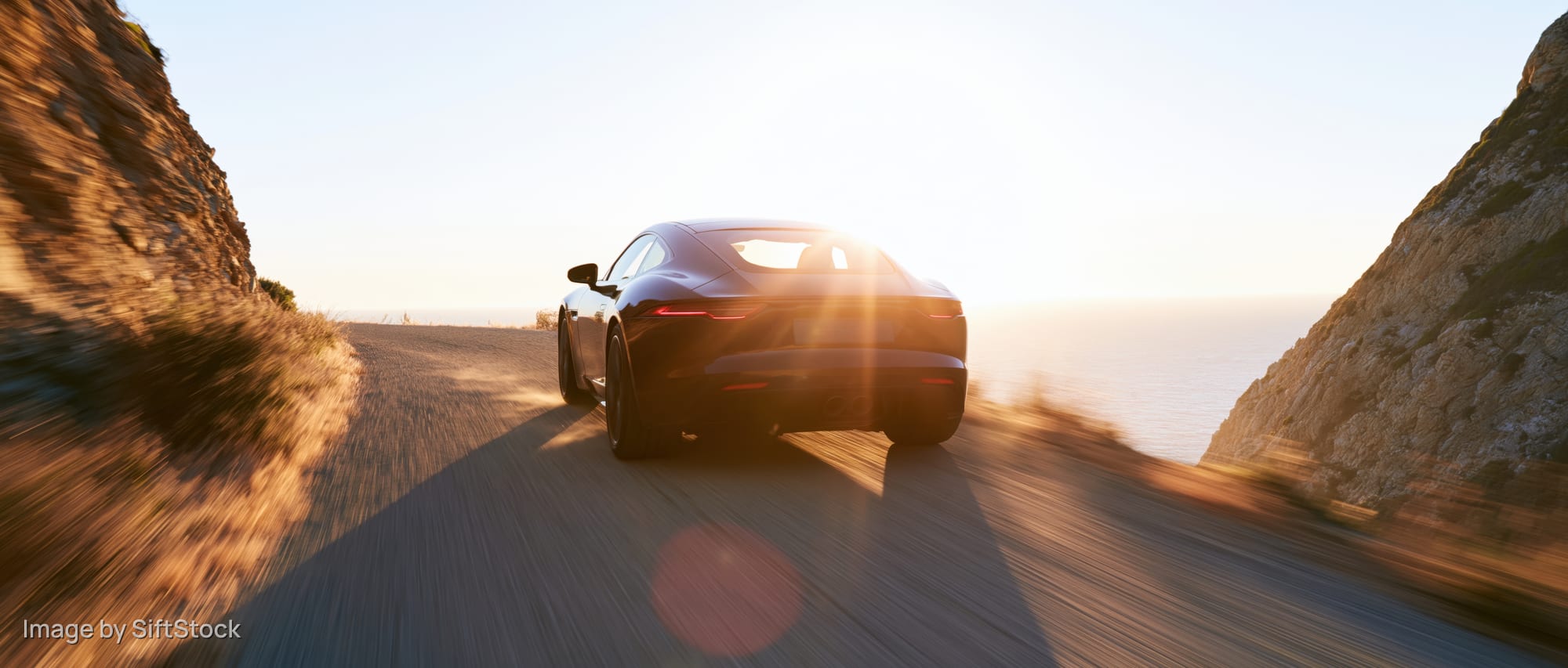 Black sports coupe driving away on narrow coastal road at sunset
