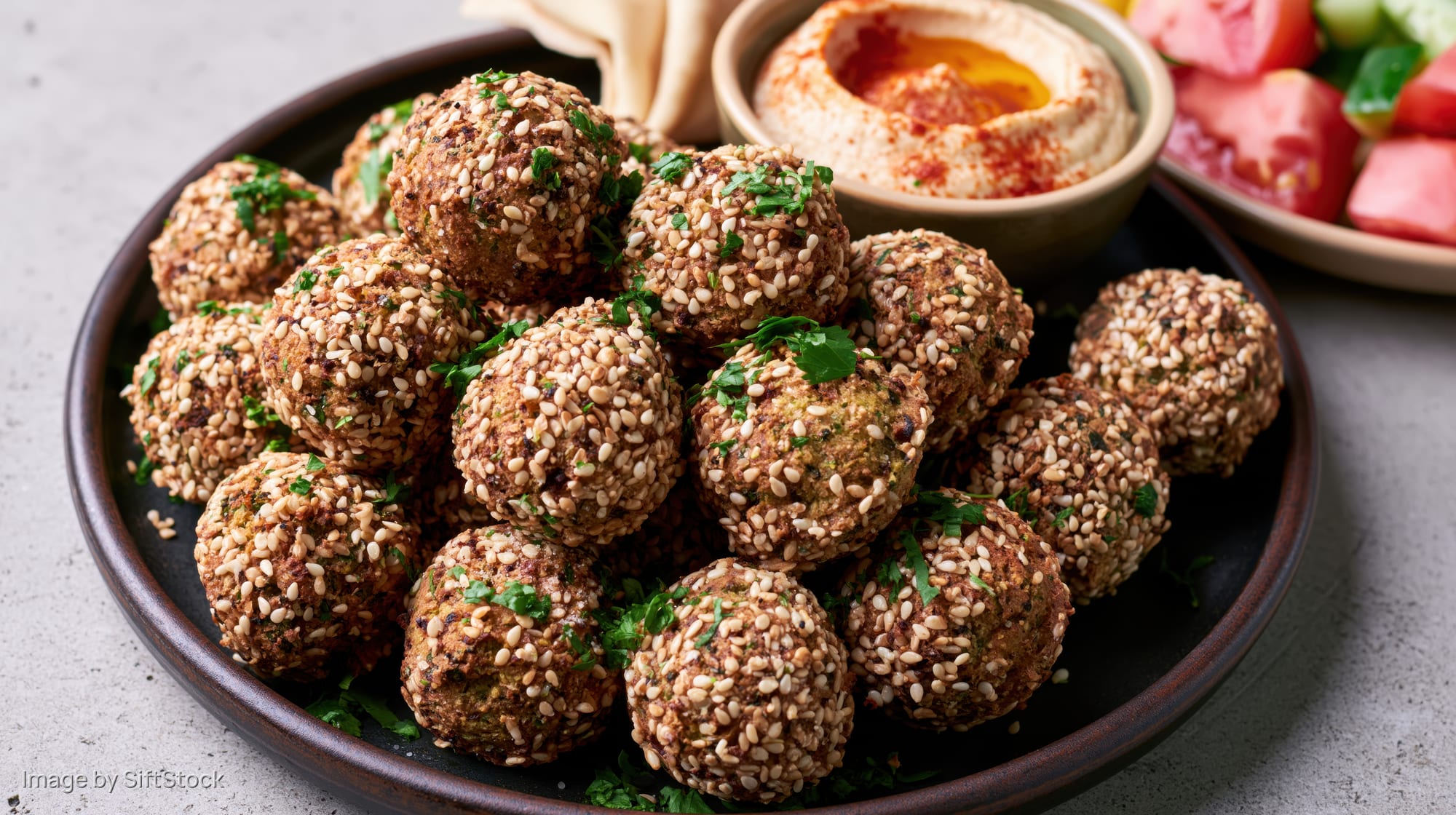 Golden-brown falafel balls coated in sesame seeds on a dark ceramic plate with hummus and salad beside