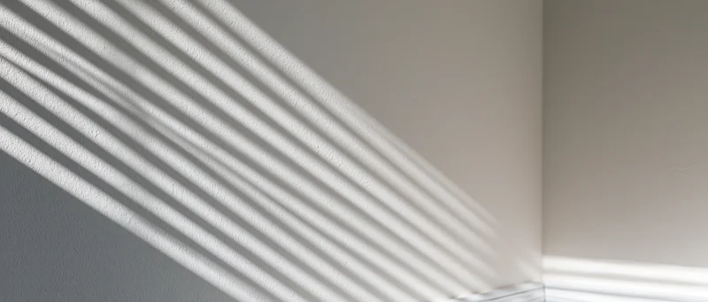 Diagonal light casting parallel shadow stripes across a white interior wall corner