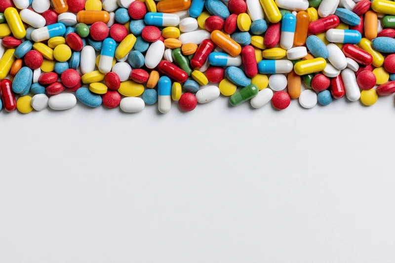 Assorted pharmaceutical pills and capsules scattered across white surface with copyspace below