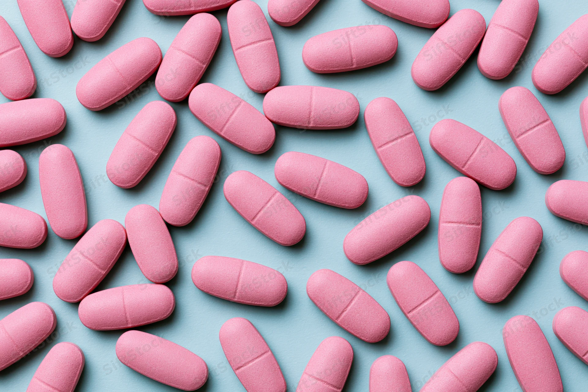Pink oval caplet tablets scattered randomly on a light blue surface in a flat lay top view