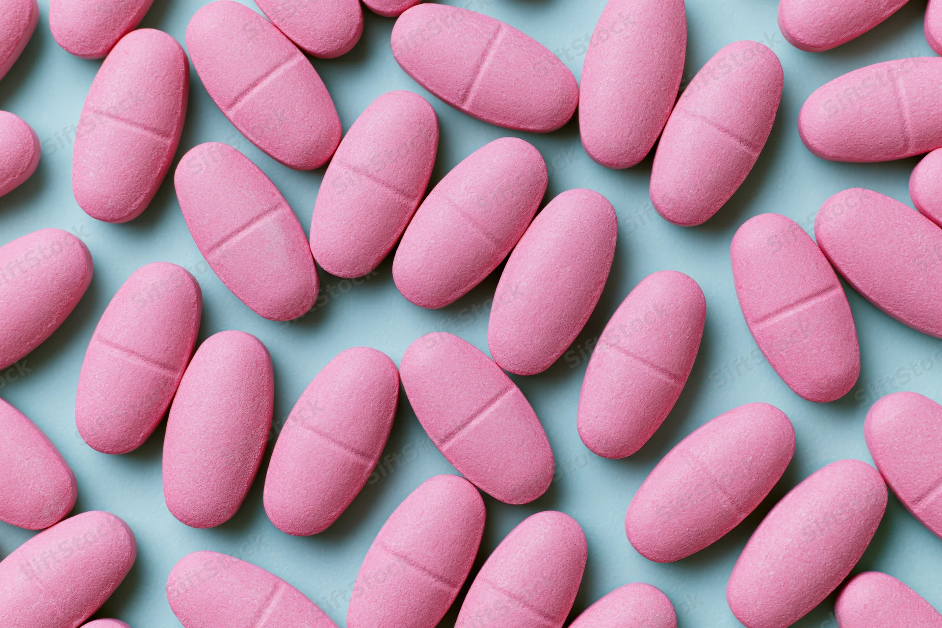 Scattered matte pink oval caplet tablets on a light blue surface